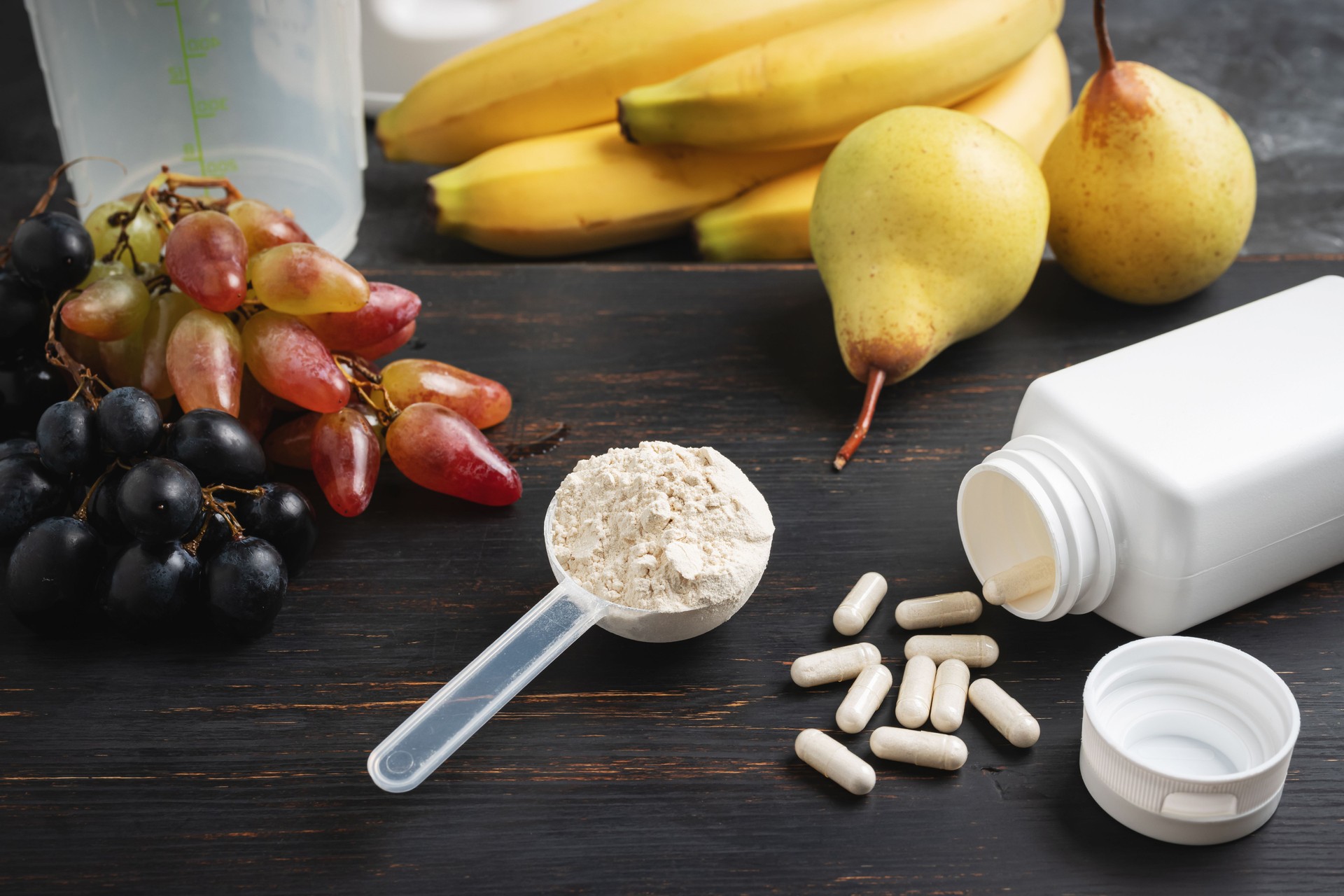 Scoop of whey or soy protein powder, white capsules of amino acids, vitamins, creatine, bodybuilding food supplements and sports nutrition. fruits, banana, pear and grape on a dark wooden board